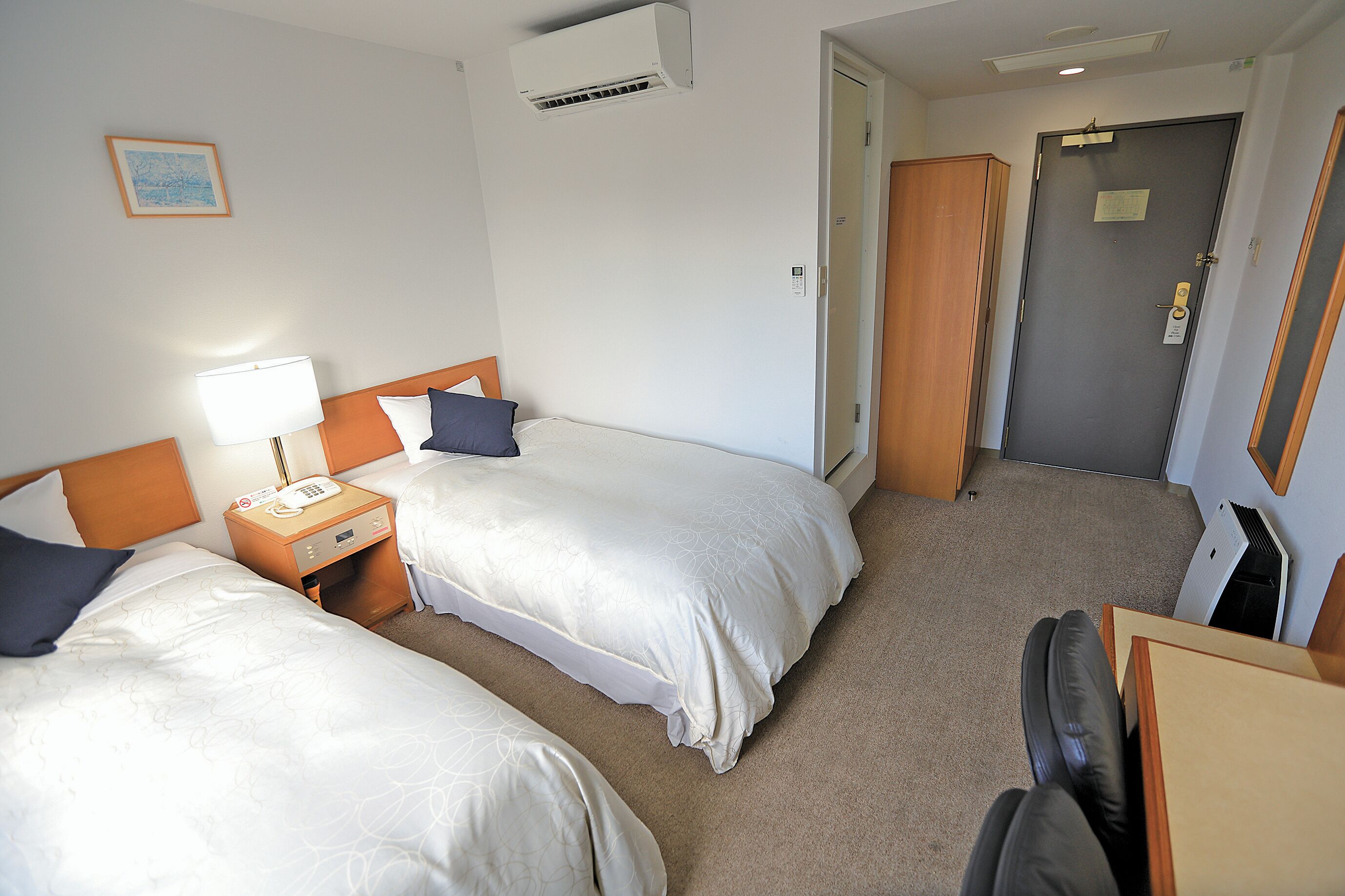 A hotel room with two beds, a bedside table with a lamp, a wooden headboard, and an air conditioning unit on the wall.