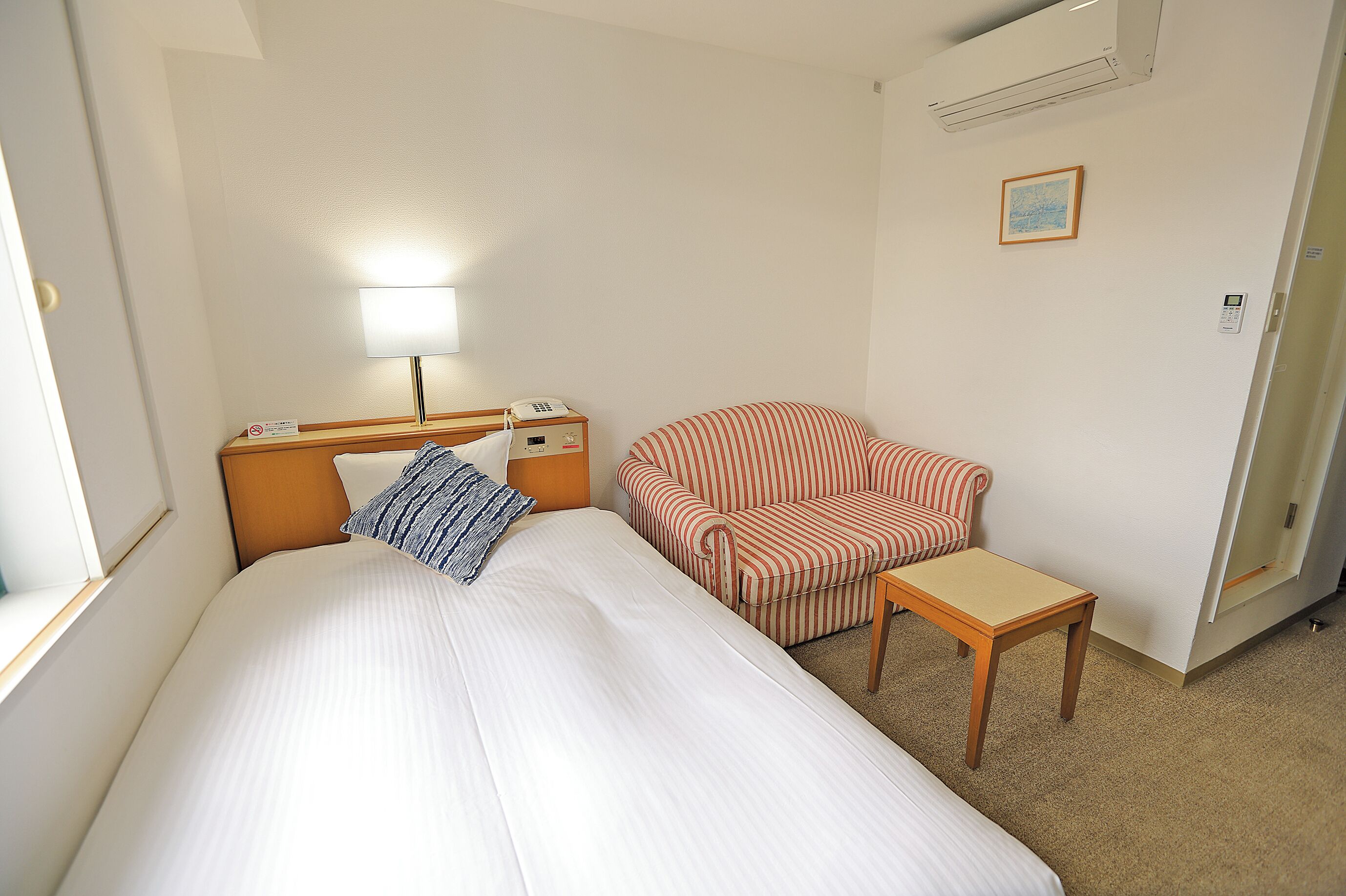 Une chambre d’hôtel avec un lit, une lampe de chevet, un téléphone, un fauteuil à rayures et une petite table.