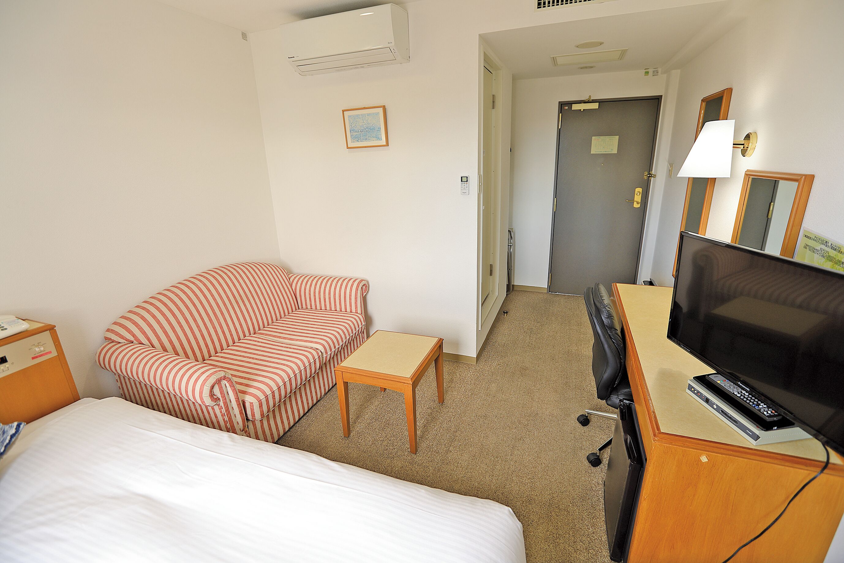 Habitación de hotel con cama, sofá a rayas, una mesita, escritorio con televisor y puerta que da a otra habitación.