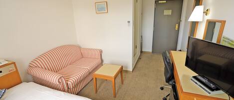 Habitación de hotel con cama, sofá a rayas, una mesita, escritorio con televisor y puerta que da a otra habitación.