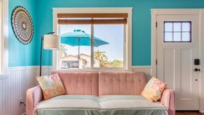 A room with a pink sofa, a window with a blue umbrella, a white door, and a wall with a decorative plate.