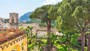 A historic building with a tower, surrounded by lush greenery and a mountainous backdrop.