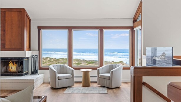 A room with a fireplace, two armchairs, a round table, and large windows with a view of the ocean.