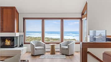 A room with a fireplace, two armchairs, a round table, and large windows with a view of the ocean.