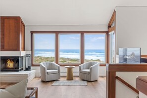 A room with a fireplace, two armchairs, a round table, and large windows with a view of the ocean. - Dunes Sea Haven: Ocean Front 3br 3ba (Moss Landing)