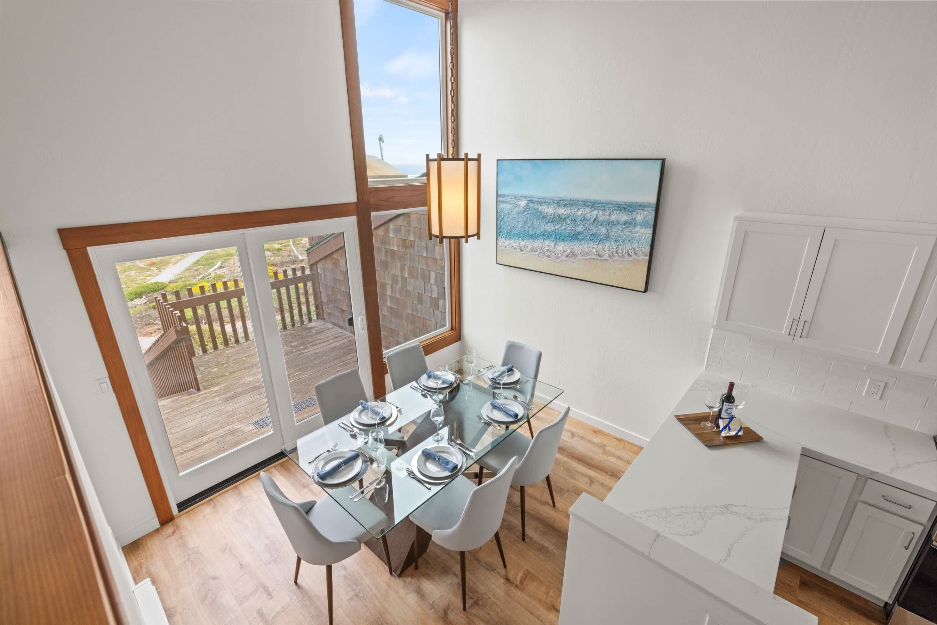 A dining area with a glass table, white chairs, and a view of the beach through a large window.