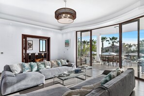 A modern living room with a grey sectional sofa, a glass coffee table, and a dining area with a wooden table and chairs. - Chateau Moderne Luxury Villa in Paphos with private pool and gardens (St Georges Peyia)