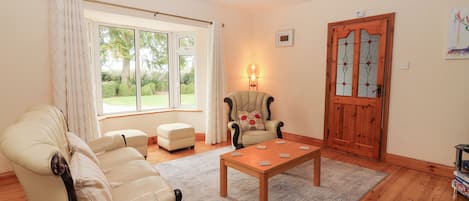 Sala de estar com sofá bege, mesa de centro de madeira e uma poltrona com almofada. Há uma janela com cortinas, uma porta com painéis de vidro e um abajur na parede.