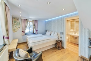 A modern bedroom with two beds, a desk, a chair, a small stool, a window with curtains, and a bathroom with a sink and mirror. - Villa Anna Kitzbühel design apartments (Kitzbuhel)