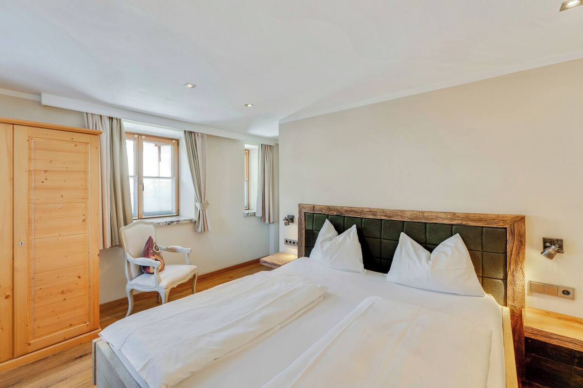 A bedroom with a large bed, a wooden wardrobe, a chair, and a window with curtains.