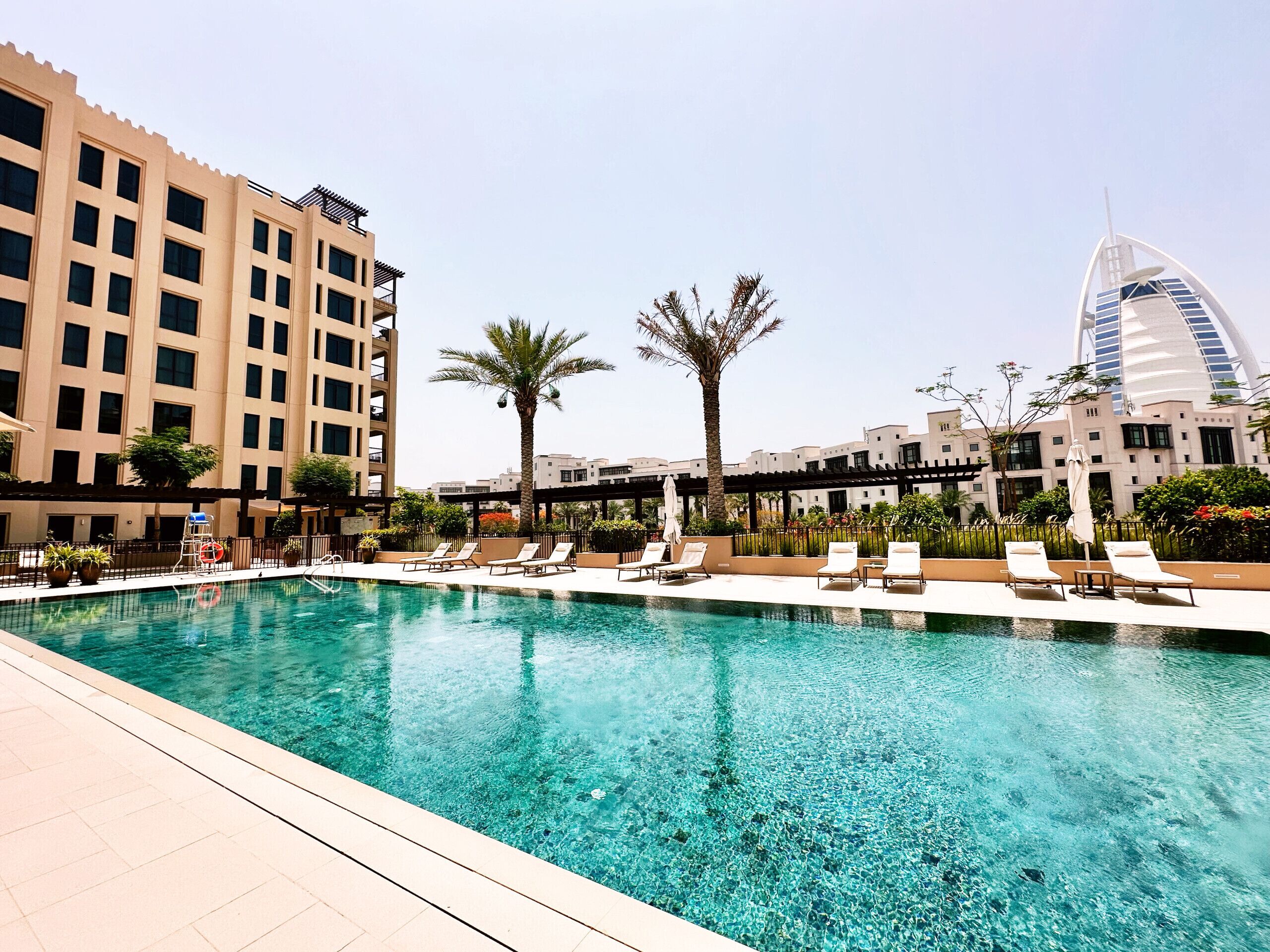 Piscina com espreguiçadeiras e palmeiras, cercada por edifícios e uma estrutura moderna imponente ao fundo.