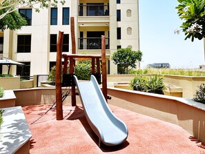 Ein Spielplatz mit Rutsche und Klettergerüst, umgeben von einer gepflegten Grünanlage mit Pflanzen und einem Gebäude im Hintergrund.