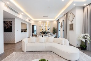 A modern living room with a curved sectional sofa, a dining area with a chandelier, and a large abstract painting on the wall.