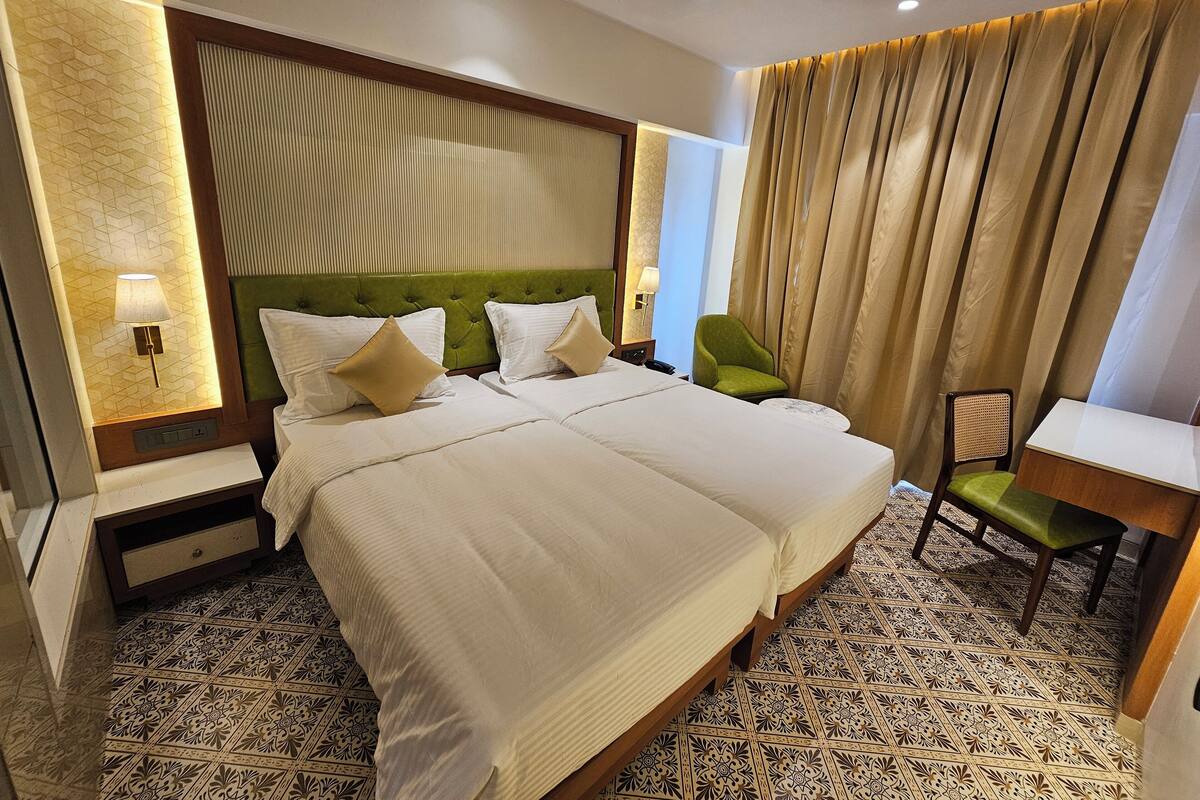 Habitación de hotel con dos camas, un cabecero tapizado en verde, un sillón verde, un escritorio pequeño y baldosas de piso estampadas.