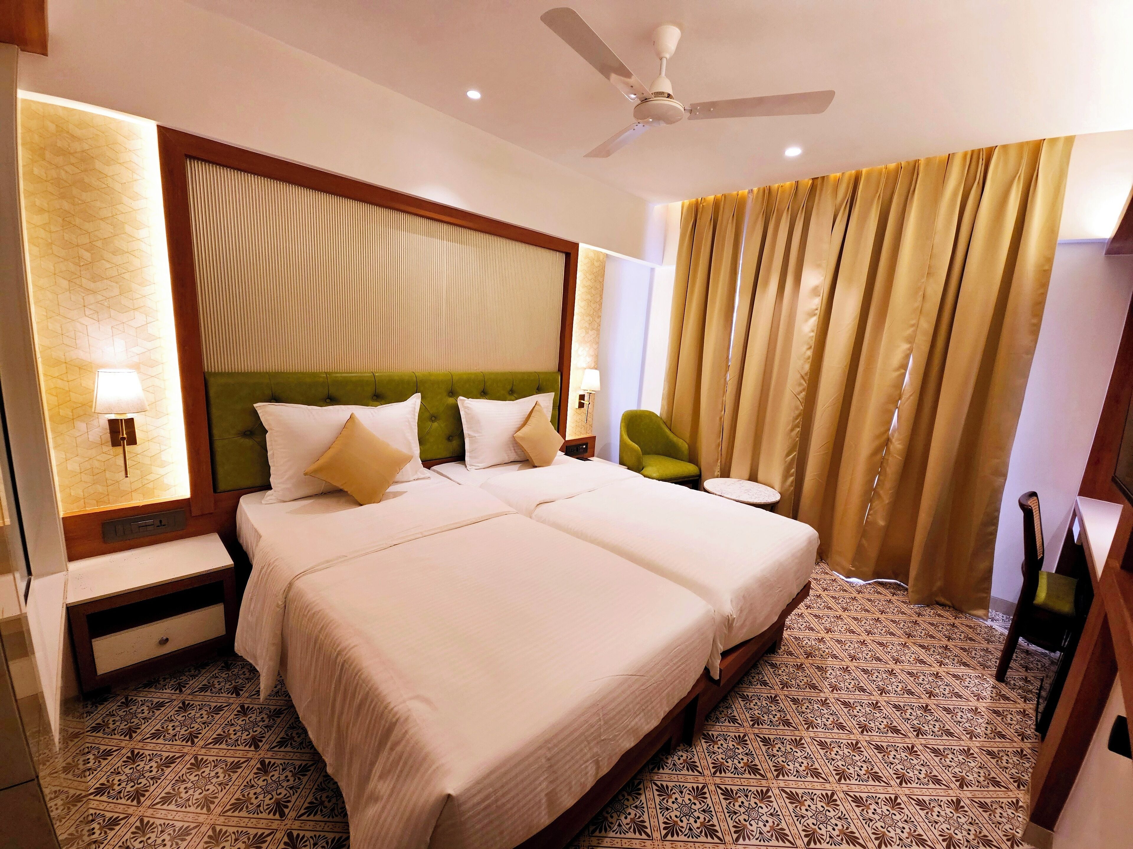 A hotel room with two beds, a green headboard, a ceiling fan, and patterned floor tiles.