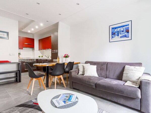 A modern living room with a grey sofa, a dining table with chairs, and a kitchen in the background.