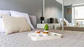 Un lit bien fait, sur lequel repose un plateau avec deux tasses et une petite plante, une table de chevet avec une lampe, et un grand miroir qui reflète la pièce.