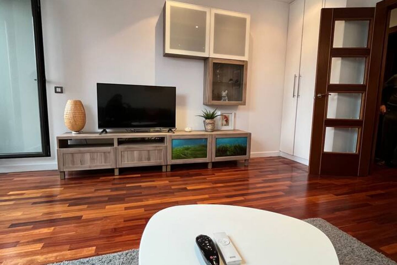 Una sala de estar con un televisor de pantalla plana, un mueble de madera para entretenimiento y una mesa de centro blanca.