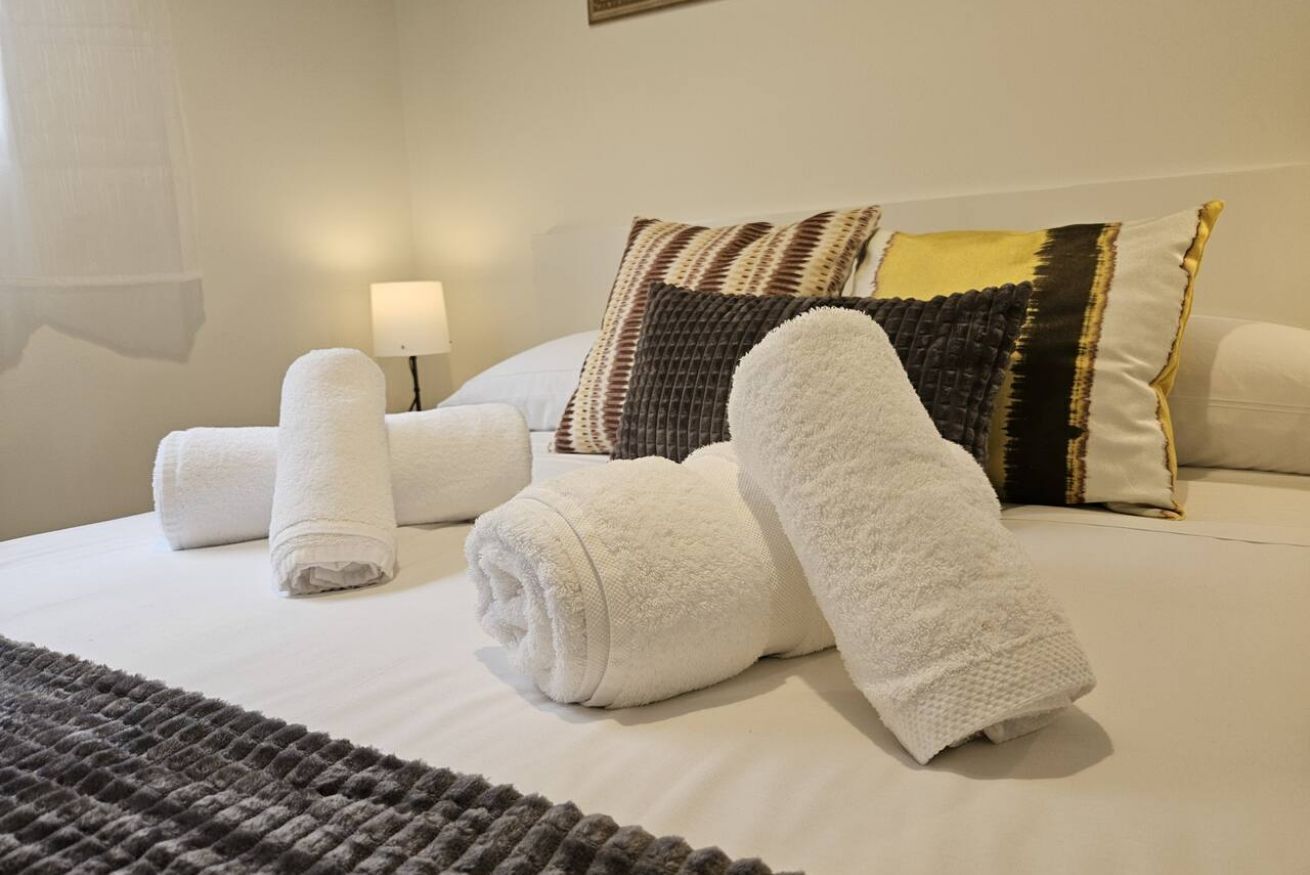 A neatly made bed with rolled white towels, a lamp, and patterned pillows.