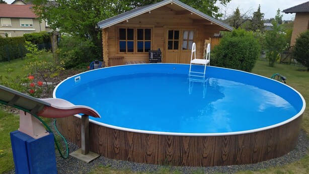 Une piscine ronde hors sol, dotée d’une terrasse en bois et d’une échelle, entourée d’un jardin et d’une maison en bois.