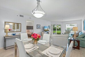 A dining area with a glass table, white chairs, and a chandelier. There is a sofa and a television in the background.