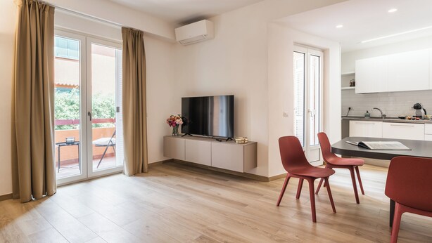 Una sala de estar moderna con un televisor de pantalla plana, una zona de comedor con sillas rojas y una cocina con armarios blancos.