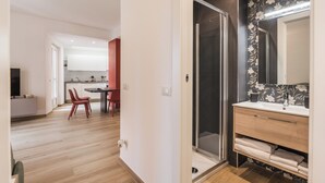 Cocina moderna con armarios blancos, comedor con sillas rojas y baño con ducha de vidrio y papel tapiz floral.