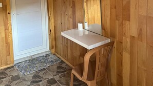 A small room with wooden walls, a white door, a mounted TV, a white table, a plastic chair, and a stone-patterned floor.