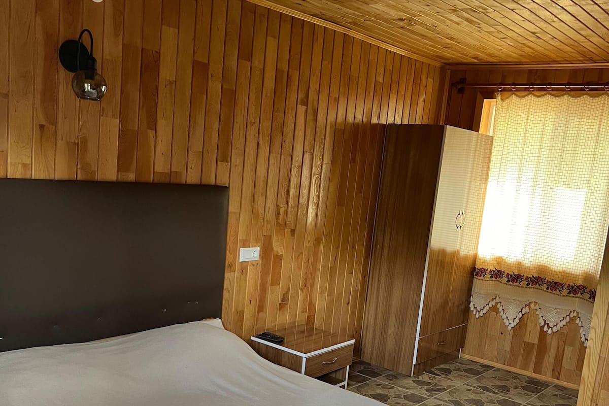 A wooden-paneled room with a bed, a nightstand, and a window with a curtain.