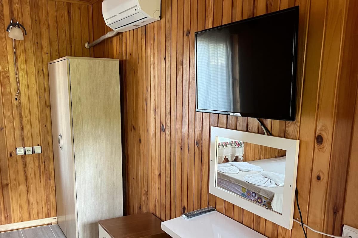 A room with wooden panel walls, a white TV mounted on the wall, a small stool, a white shelf, and a wooden wardrobe.