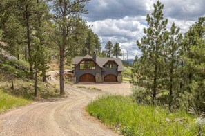 A rustic cabin with a dirt driveway, surrounded by pine trees, and a mountain range in the background. - Castle in the Sky- Overlooking Rapid City (Rapid City)