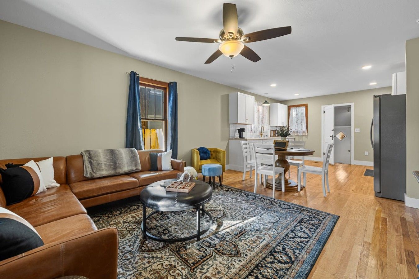 A living room with a sofa, coffee table, and a patterned rug. There is a ceiling fan and a kitchen area in the background.