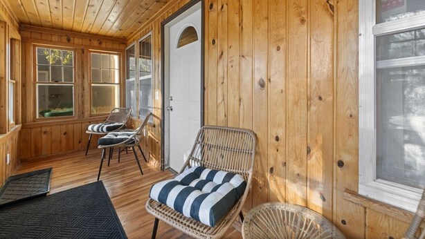 Een houten blokhut van binnen met een veranda, rieten stoelen, een met kussens beklede bank en een deur.