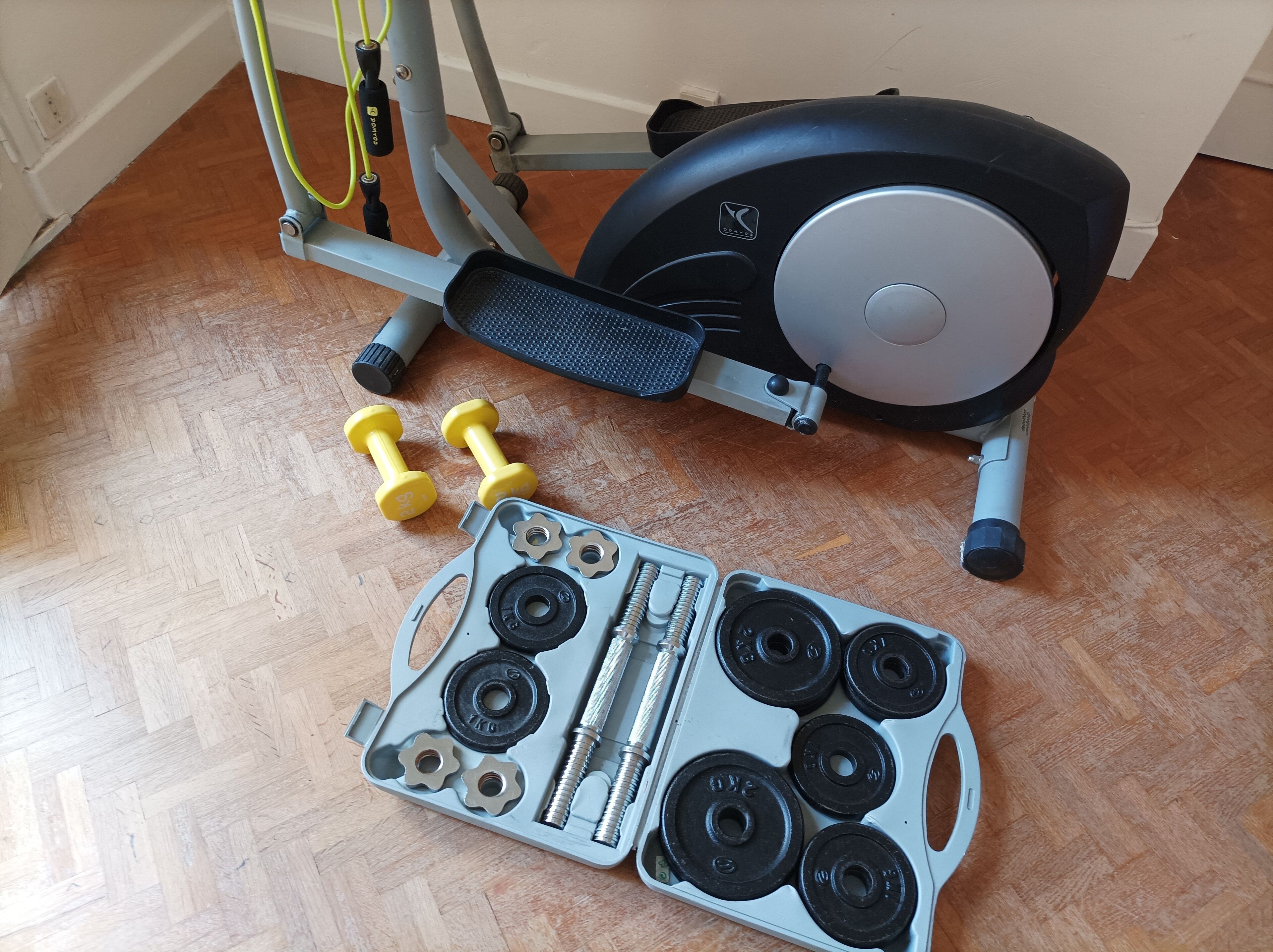 A stationary exercise bike, two yellow dumbbells, and a weight plate set on a wooden floor.