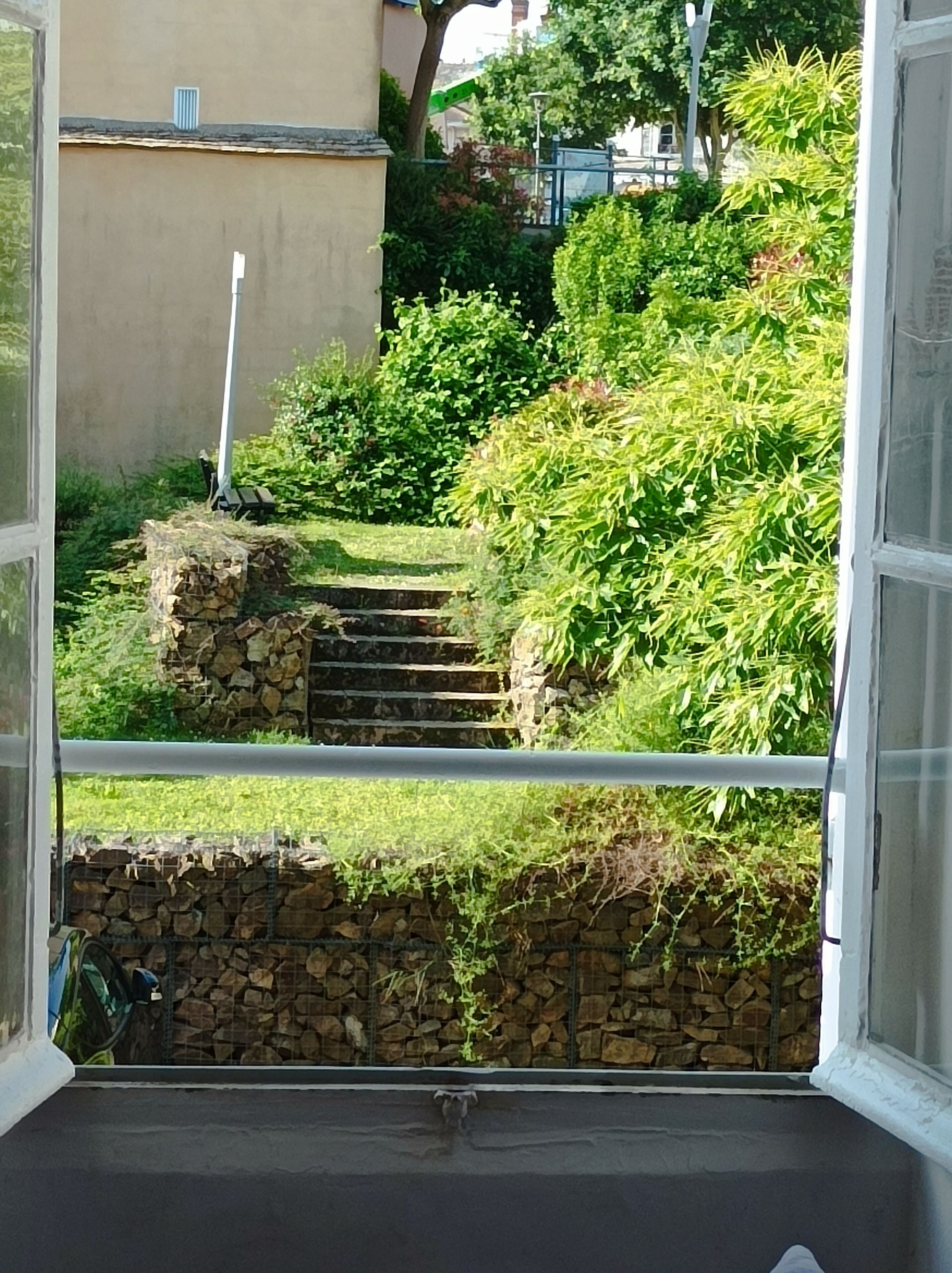 A view from a window overlooking a garden with stone steps leading to a stone wall, surrounded by greenery.