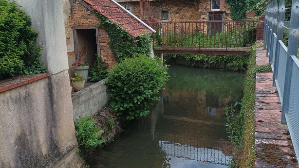 A narrow canal with a stone building on one side and a metal fence on the other. - The house by the water (Graçay)
