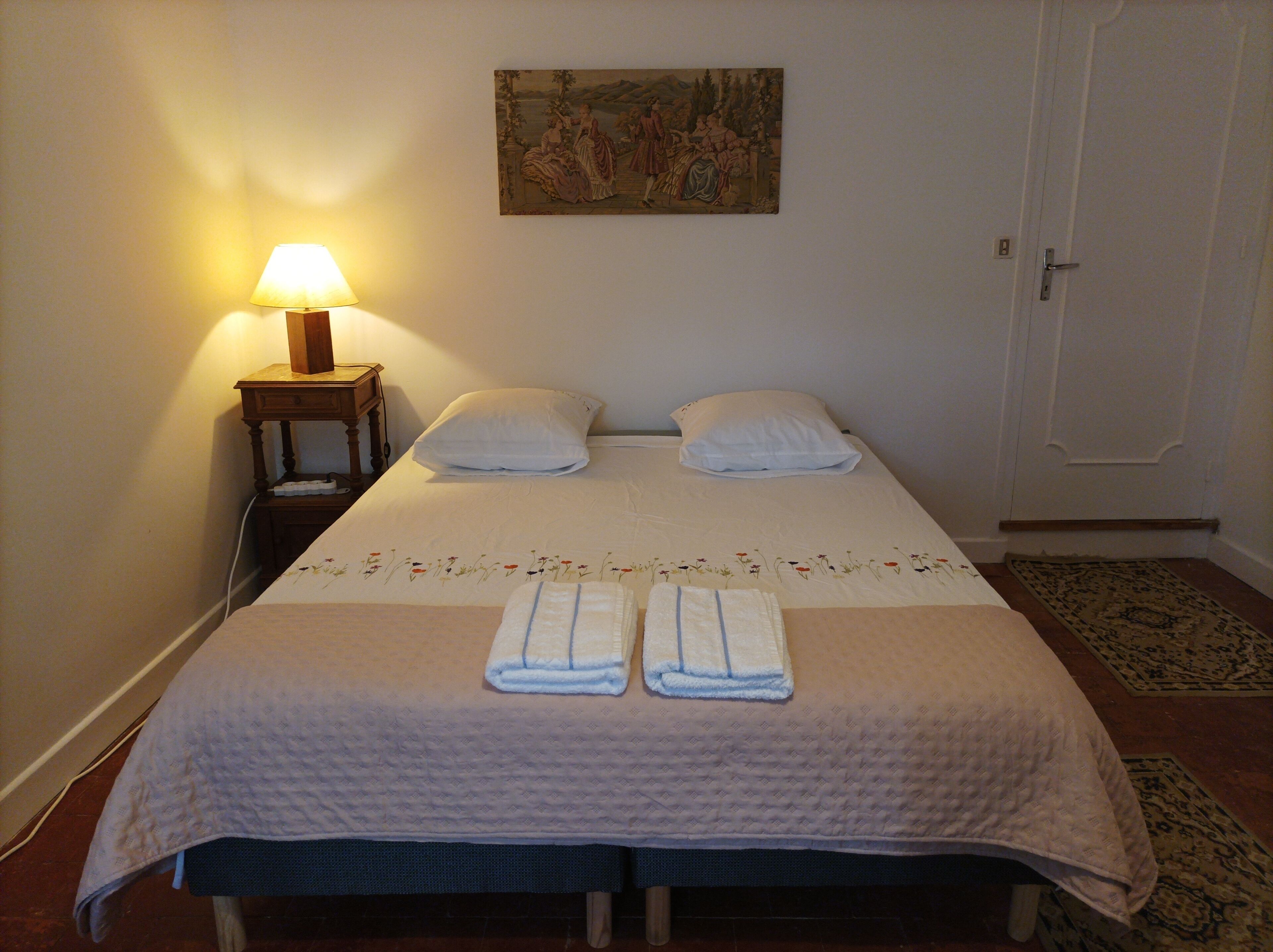 Cama bem arrumada com colcha, dois travesseiros e duas toalhas dobradas. Uma pequena mesa com abajur ao lado da cama.
