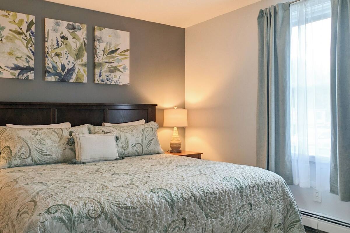 A bedroom with a large bed, a nightstand with a lamp, floral wall art, and a window with curtains.