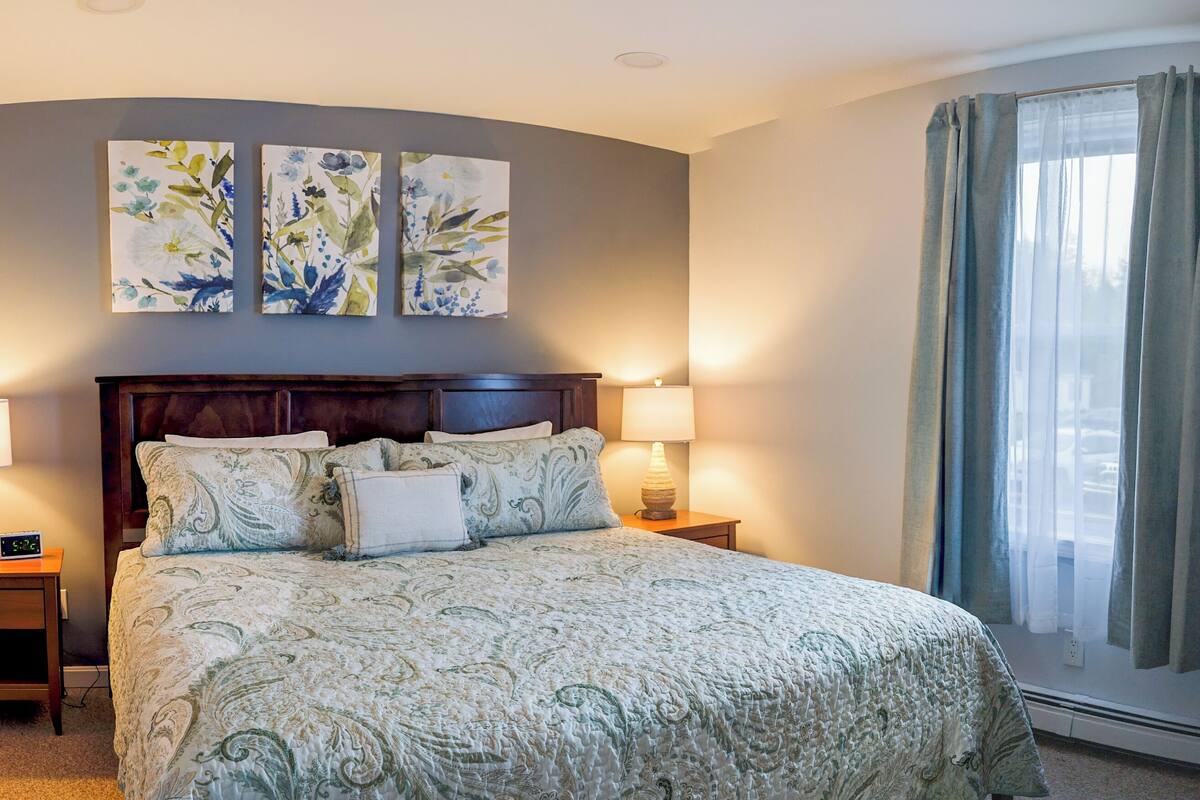 A neatly made bed with a patterned bedspread, two wall-mounted paintings, a bedside lamp, and a window with curtains.