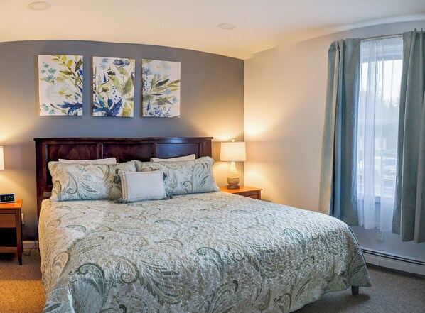 A neatly made bed with a patterned bedspread, two wall-mounted paintings, a bedside lamp, and a window with curtains.