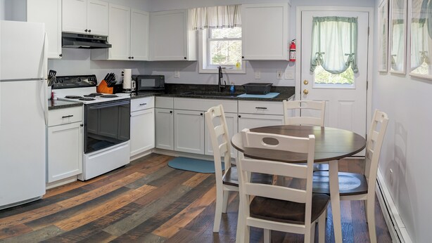 A compact kitchen with white cabinets, a stove, microwave, and a dining area with a table and chairs. - Woodland Apartments-Acadia National Park (Bernard)