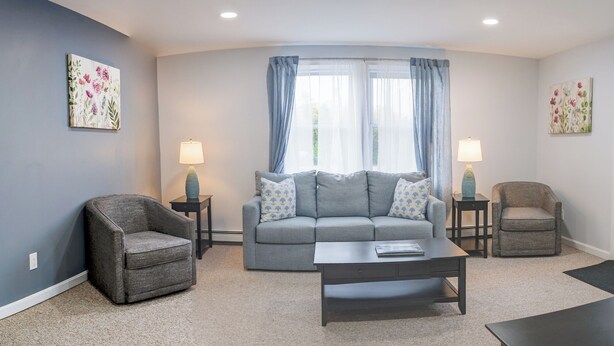 A living room with a grey sofa, two armchairs, a coffee table, and two side tables with lamps. - Woodland Apartments-Acadia National Park (Bernard)