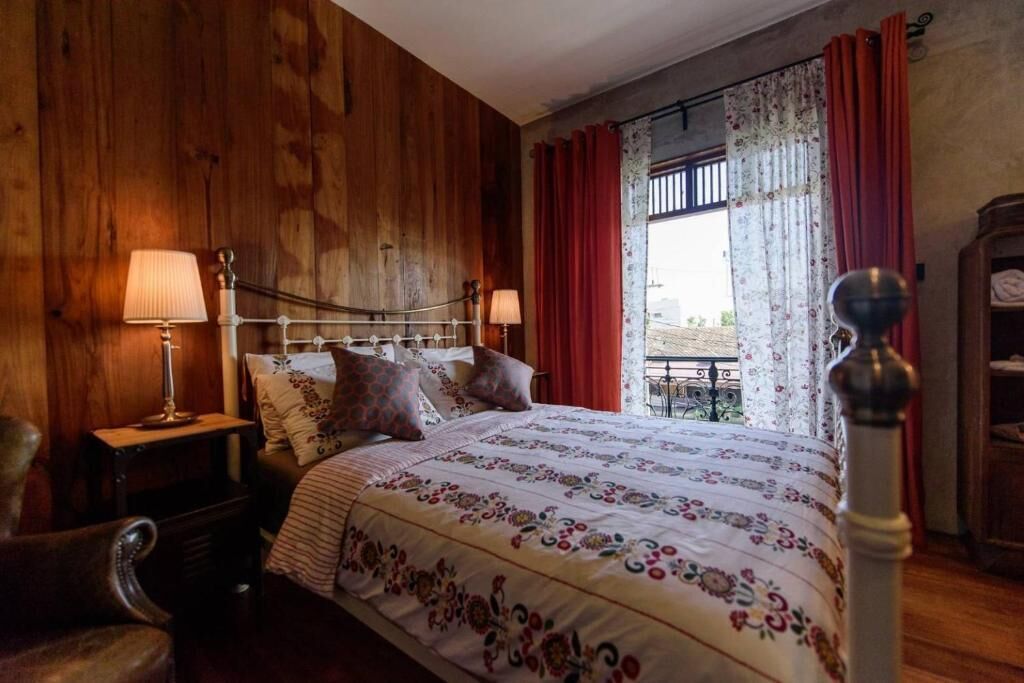 A bedroom with a wooden wall, a bed with floral bedding, two bedside lamps, and a view through a window with red curtains.