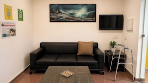 Una sala de estar con un sofá de cuero negro, una mesa de centro con un adorno en la parte superior, un cuadro sobre el sofá, un televisor en la pared y una planta en una mesita auxiliar.