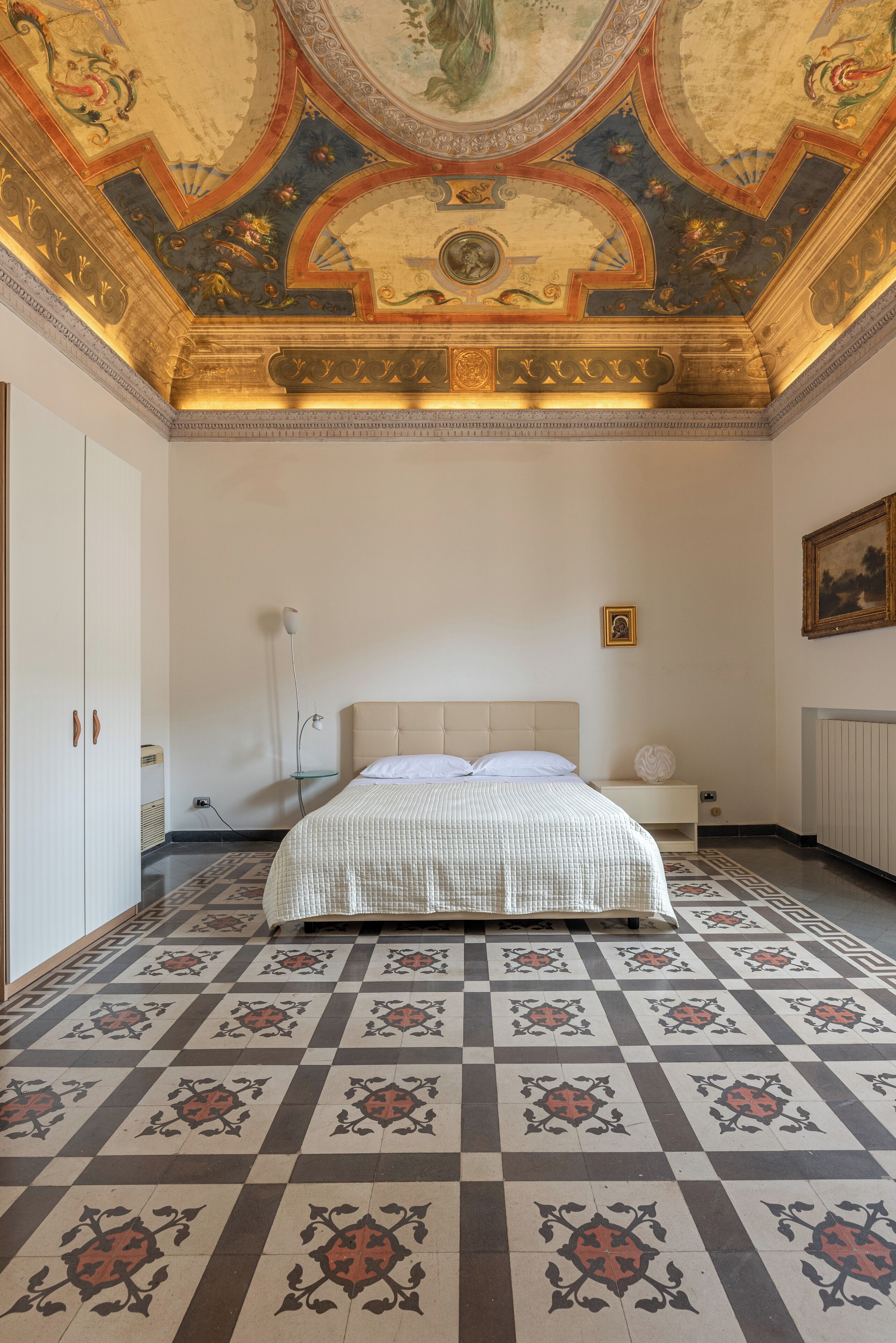 A bedroom with a checkered floor, a bed with a white headboard, a nightstand, a wall-mounted picture, and a ceiling with intricate designs.