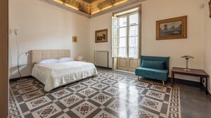 A bedroom with a bed, a blue armchair, a table, a radiator, and a window with curtains. - Amari Elegant Apartment by Wonderful Italy (Palermo)