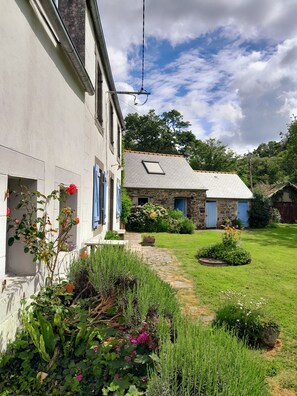 A white building with blue shutters, a stone house with a tiled roof, and a well-maintained garden with various plants and flowers. - Beautiful country house at the entrance to the Crozon peninsula (Rosnoën)