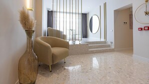 Un intérieur moderne avec un fauteuil beige, un vase en verre contenant de l’herbe des pampas, un grand vase, un miroir et un escalier menant à une autre pièce.