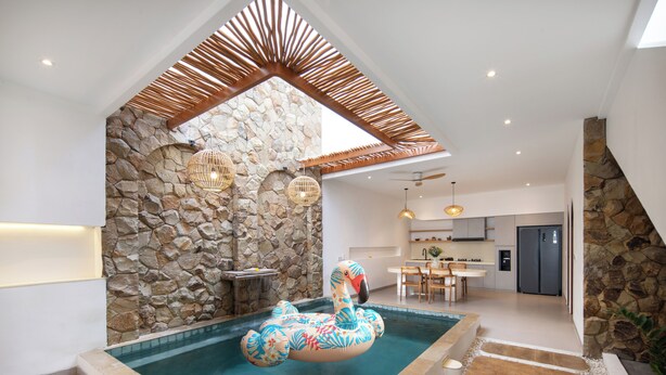 A swimming pool with a decorative flamingo float, stone walls, a wooden ceiling, and a kitchen area in the background. - Onnea Villa by Lead (Canggu)