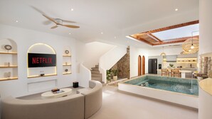 A modern interior with a curved seating area, a Netflix sign, a pool, and a kitchen in the background. - Onnea Villa by Lead (Canggu)